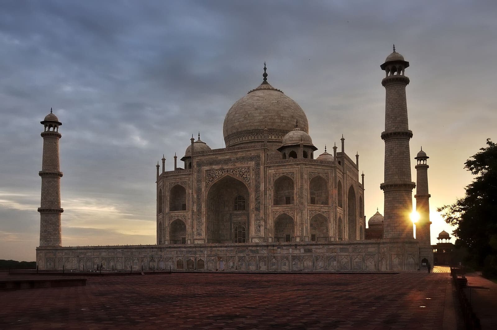 The white marble main mausoleum of the Taj Mahal and one of its minarets at sunrise, under a dramatic sky