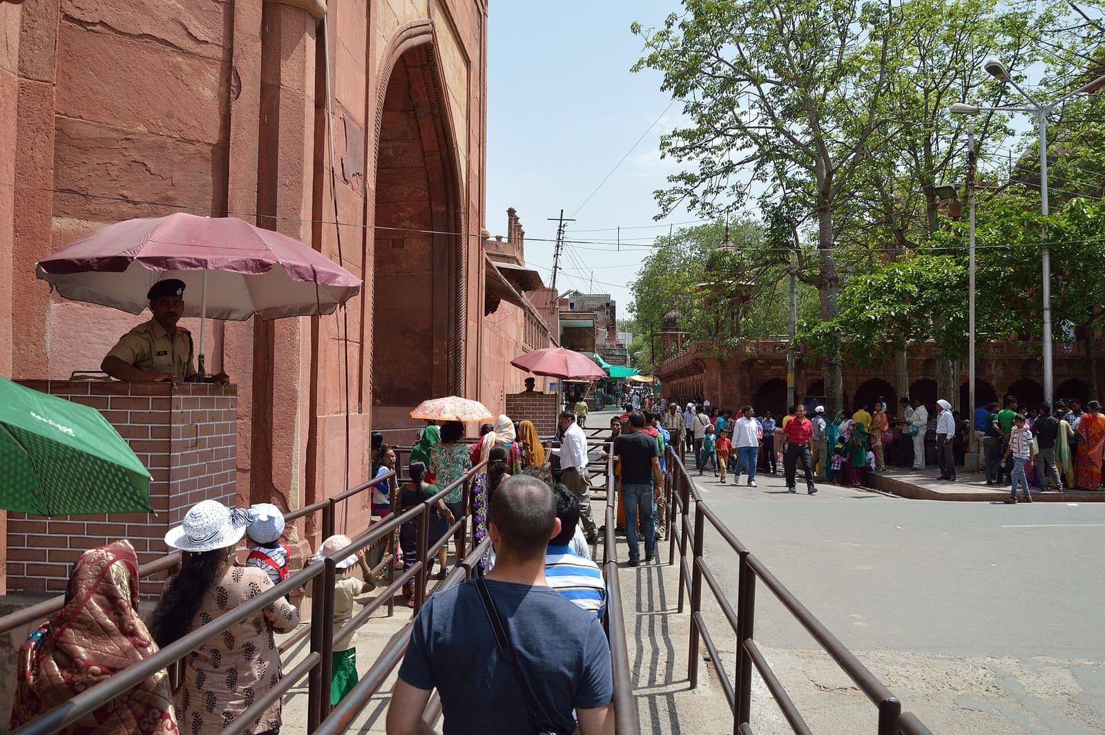 Visitors queueing between railings beside the red sandstone wall of the Taj Mahal’s West Gate, with a police post under an umbrella