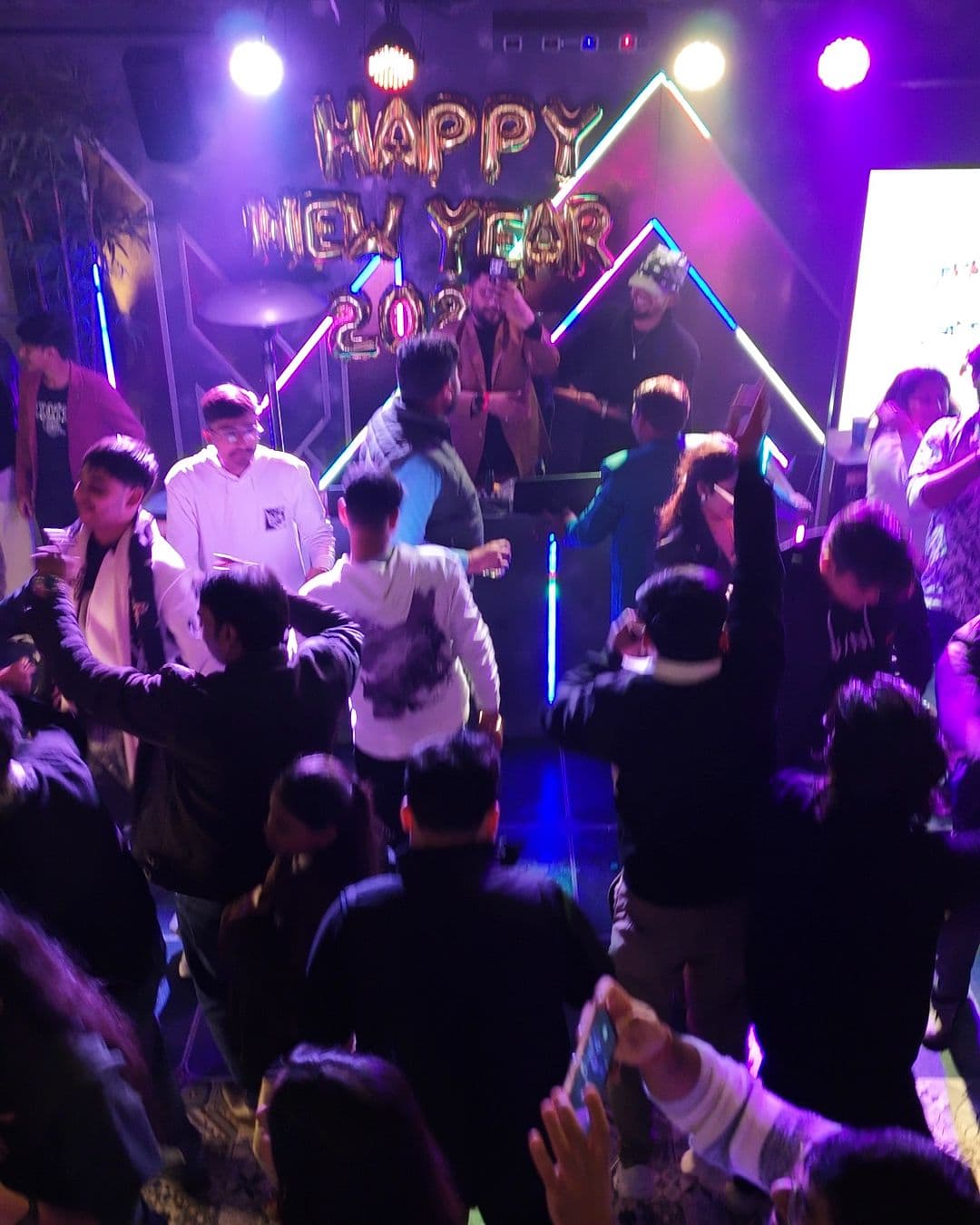 A full dance floor in violet and blue light on New Year’s Eve 2025, hands in the air, the HAPPY NEW YEAR balloons behind the DJ