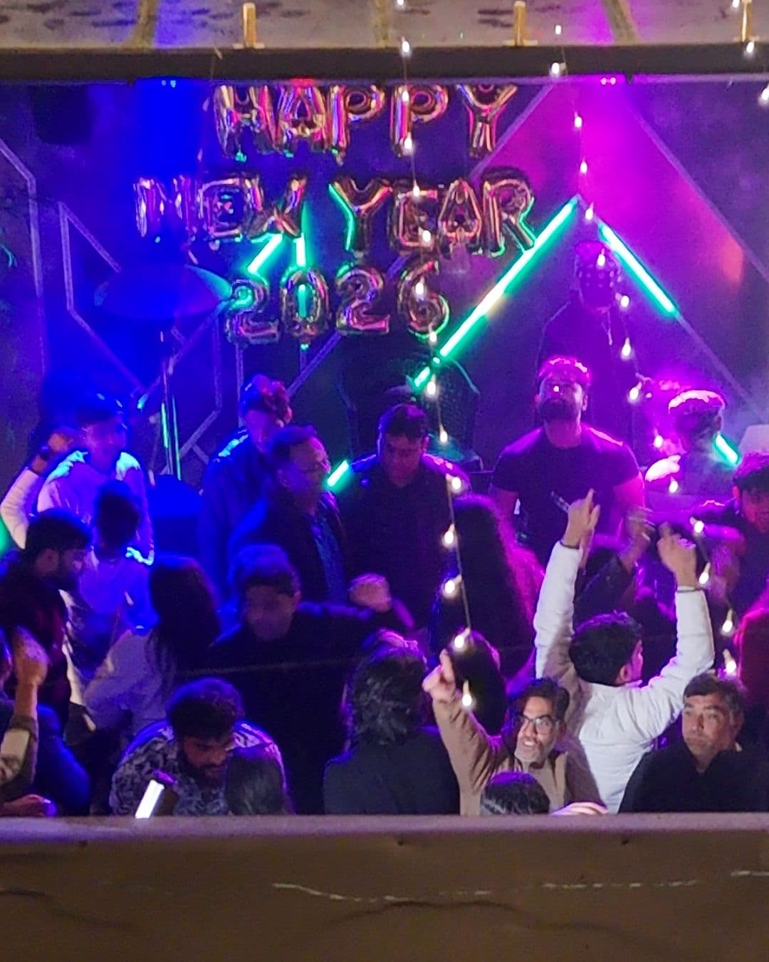Guests dancing with their hands up under gold HAPPY NEW YEAR 2026 balloons and neon light, seen from outside the roof